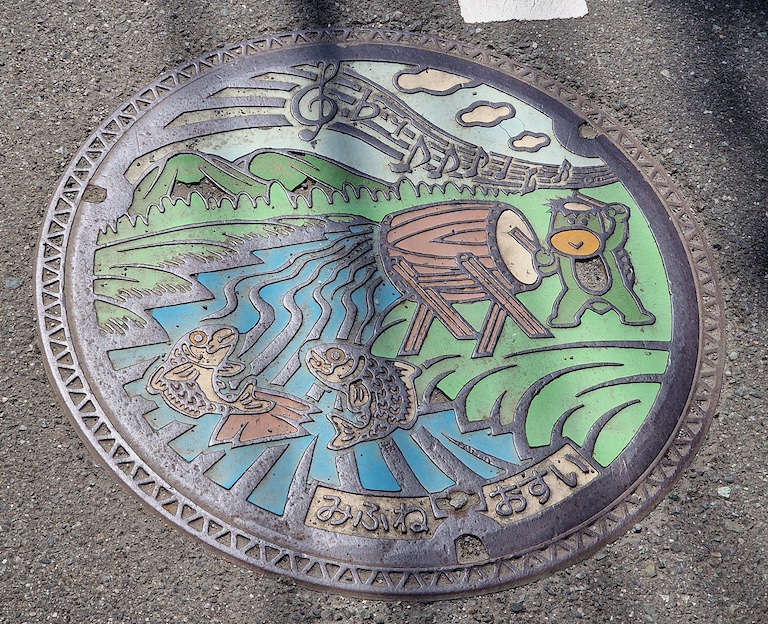 A manhole cover depicting a Kappa banging a drum for two dancing fish