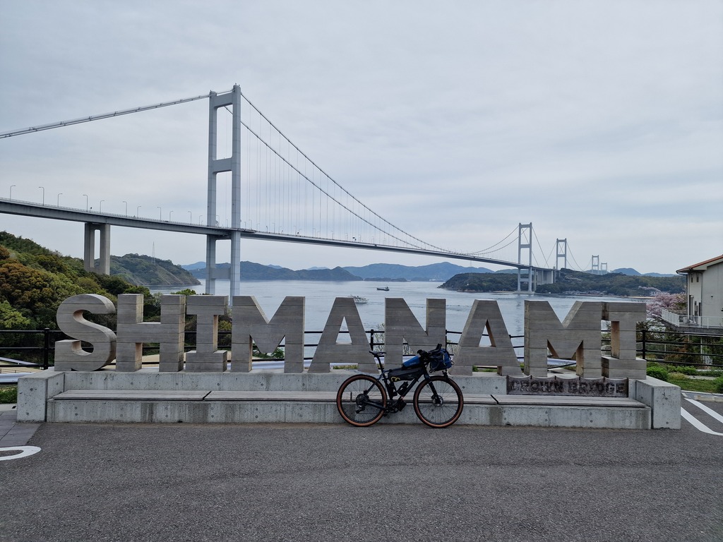 My bicycle leaning on a sign spelling Shimanami in big wooden letters. In the background you can see the Kurushima Strait bridge.